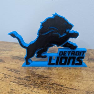 May include: A black and blue 3D-printed Detroit Lions logo. The logo features a leaping lion over the words "DETROIT LIONS" in blue. The lion is black with a blue outline, and the base is blue.