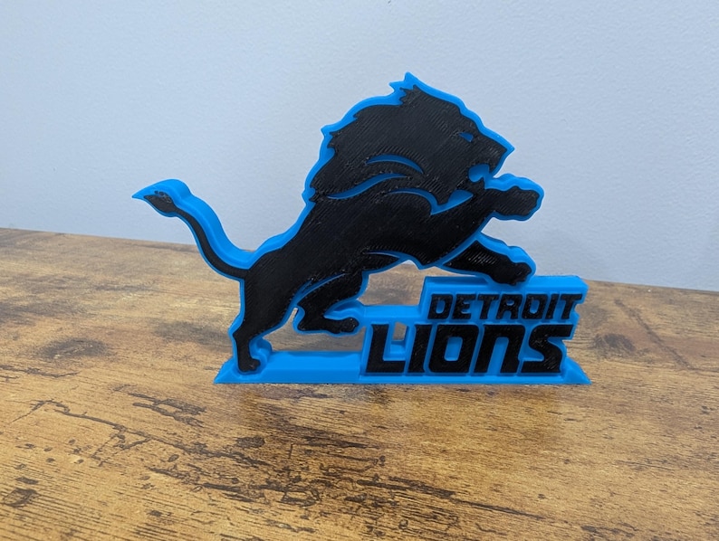 May include: A black and blue 3D-printed Detroit Lions logo. The logo features a leaping lion over the words "DETROIT LIONS" in blue. The lion is black with a blue outline, and the base is blue.