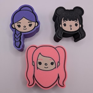 Puede incluir: Tres accesorios coloridos con forma de caras de personajes de dibujos animados. Uno es morado con un peinado trenzado, otro es negro con dos moños y el último es rosa con coletas. Cada uno tiene una cara sonriente con contornos negros.
