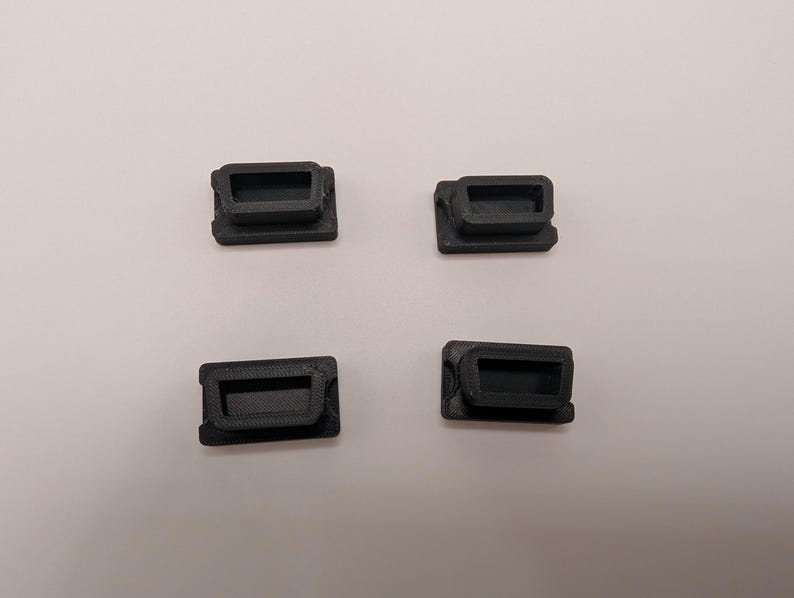 Puede incluir: Cuatro peque&ntilde;os componentes rectangulares negros de pl&aacute;stico est&aacute;n dispuestos sobre una superficie blanca. Cada componente tiene una abertura rectangular empotrada y un marco ligeramente elevado. Los componentes parecen estar impresos en 3D.
