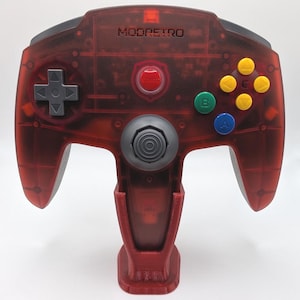 Pode incluir: Um comando de videojogos vermelho translúcido com o texto MODRETRO impresso na parte frontal. Possui um pad direcional cinzento, um joystick analógico central cinzento, um botão start vermelho e quatro botões de ação coloridos com A, B e setas amarelas.