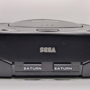 Puede incluir: Una consola de videojuegos Sega Saturn negra. La consola tiene la palabra "SEGA" en letras plateadas en la parte delantera. Debajo del logotipo hay dos ranuras con la etiqueta "SATURN". La consola es rectangular con una parte superior curvada.
