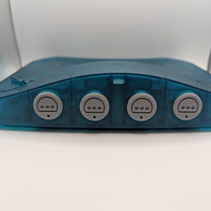 Puede incluir: Una consola de videojuegos de color turquesa transparente con cuatro puertos para mandos. La consola tiene una forma rectangular redondeada con una parte superior ligeramente curvada. Los puertos para mandos son grises y circulares, con tres pequeños agujeros.