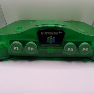 Puede incluir: Una consola de juegos Nintendo 64 verde transparente. La consola tiene cuatro puertos de controlador etiquetados P1 a P4. El logotipo de Nintendo 64 es visible en la parte delantera. La consola está sobre una superficie blanca.