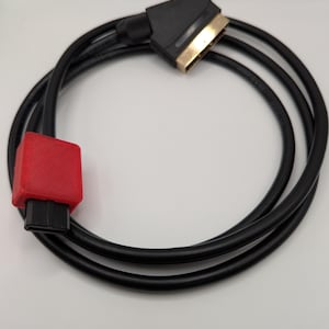 Pode incluir: Um cabo AV preto com um conector SCART dourado numa extremidade e um conector preto dentro de uma caixa de plástico vermelho texturizado na outra. O cabo está enrolado numa forma circular, exibindo o fio preto e os conectores.