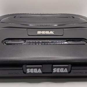 Puede incluir: Una consola Sega Genesis negra, un sistema de videojuegos cl&aacute;sico. La consola tiene forma rectangular con una parte superior ligeramente curvada. El logotipo de Sega es visible en la parte delantera y superior. La consola tiene dos puertos de controlador y una ranura para cartuchos.