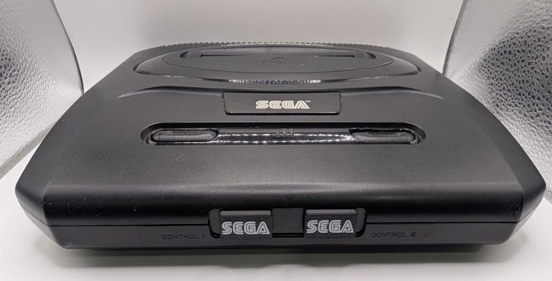 Puede incluir: Una consola Sega Genesis negra, un sistema de videojuegos cl&aacute;sico. La consola tiene forma rectangular con una parte superior ligeramente curvada. El logotipo de Sega es visible en la parte delantera y superior. La consola tiene dos puertos de controlador y una ranura para cartuchos.