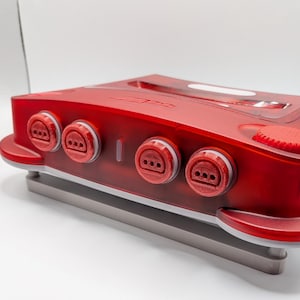 May include: A translucent red video game console featuring four controller ports on the front panel, a circular red power button on the top right, and a grey base. The console has a sleek, curved design with a visible cartridge slot on the top.