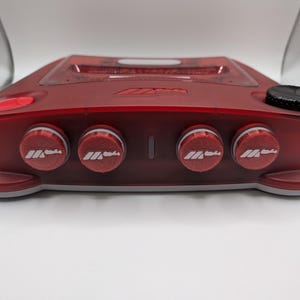 Könnte beinhalten: Ein durchscheinendes rotes elektronisches Gerät mit vier runden Drehreglern auf der Vorderseite, die jeweils mit einem weißen Logo versehen sind. Die Oberseite enthält ein zentrales Display, einen roten Knopf und ein schwarzes Einstellrad.