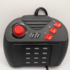 May include: Black Atari controller with a red joystick and red A, B, and C buttons. The controller has a numeric keypad and pause and option buttons. The Atari logo is visible on the top.