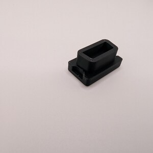 Puede incluir: Un peque&ntilde;o componente de pl&aacute;stico negro, rectangular, con una abertura rectangular hueca. El componente tiene una base con un peque&ntilde;o agujero en un lado. El objeto est&aacute; sobre un fondo liso y claro.