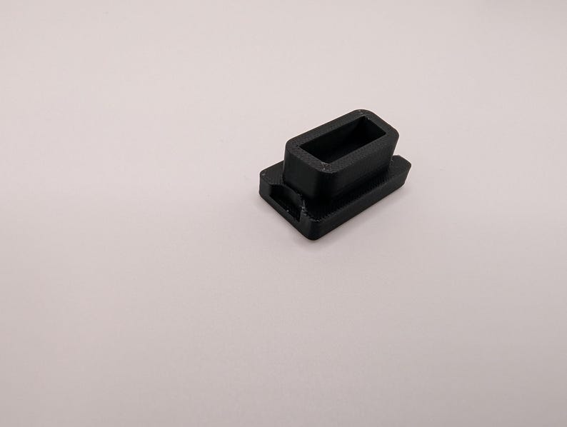 Puede incluir: Un peque&ntilde;o componente de pl&aacute;stico negro, rectangular, con una abertura rectangular hueca. El componente tiene una base con un peque&ntilde;o agujero en un lado. El objeto est&aacute; sobre un fondo liso y claro.