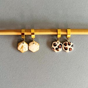 May include: Two pairs of hexagon-shaped earrings. One pair features a white base with gold flecks, the other has a white base with brown and black leopard print. Both pairs have gold-colored hoops.