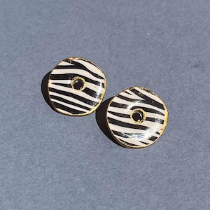 May include: A pair of round stud earrings with a zebra stripe pattern in black and white. The earrings have a gold-colored border and a small black center. The earrings are displayed on a gray surface.