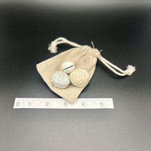 May include: Three decorative, textured spheres in shades of gray and beige rest on a small, beige drawstring bag. One sphere has a white stripe. A ruler is placed below the bag, showing measurements in inches.