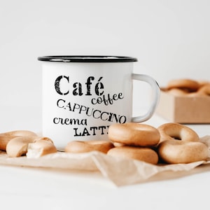 Könnte beinhalten: Eine weiße Emailletasse mit schwarzem Rand und Henkel. Die Tasse hat schwarzen Text, der "Cafe coffee Cappuccino crema Latte" lautet. Die Tasse steht auf einer braunen Papieroberfläche mit einem Stapel kleiner, runder, brauner Brötchen.