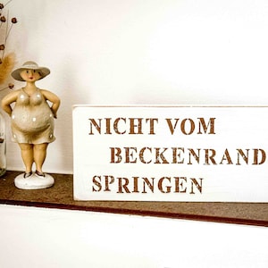 Könnte beinhalten: Ein weißes Holzschild mit braunem Text, der "NICHT VOM BECKENRAND SPRINGEN" lautet. Das Schild steht auf einem Regal mit einer kleinen Figur einer Frau mit Hut und einem gepunkteten Kleid.