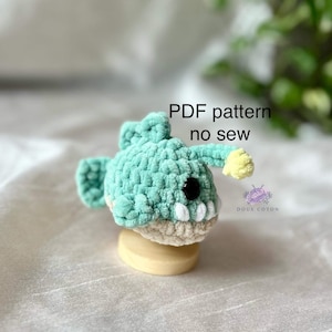 Puede incluir: Un juguete amigurumi de pez pescador de crochet con un cuerpo verde, dientes blancos y un señuelo amarillo. El juguete está sentado sobre una base de madera. El texto "PDF pattern no sew" está escrito sobre el juguete.