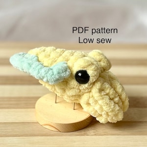May include: A handmade, plush, yellow and light blue crocheted creature with a large black eye. The text "PDF pattern Low sew" is displayed above the creature. The plush is mounted on a small wooden stand.