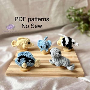 Peut inclure: Cinq petites créatures marines crochetées incluant un poisson jaune, une raie bleu clair, un poisson rayé noir et blanc, un poisson bleu et noir, et un phoque gris. Le texte indique PDF patterns No Sew et Doux Coton.