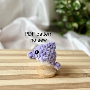 Peut inclure: Un jouet en crochet amigurumi en forme de dauphin violet avec des yeux noirs. Le jouet est sur un support en bois. Patron PDF sans couture.