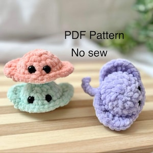 Peut inclure: Deux jouets en crochet en forme de soucoupe volante, l'un rose et l'autre bleu clair, avec des yeux noirs. Un jouet en crochet en forme de raie manta violette avec des yeux noirs est également représenté. L'image comprend le texte "PDF Pattern No sew".