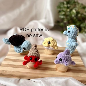 Peut inclure: Cinq animaux en peluche crochetés, dont un hippocampe, une tortue, un crabe, un dauphin et un poisson-globe. Les animaux sont présentés sur une surface en bois avec le texte "PDF patterns no sew" au-dessus.