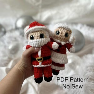 May include: Crocheted Santa Claus and Mrs. Claus dolls. The Santa doll wears a red suit with a white beard and hat, and a gold belt buckle. Mrs. Claus is in a red dress with a white collar and hat. The dolls have large, black eyes. Text on the image reads "PDF Pattern No Sew."