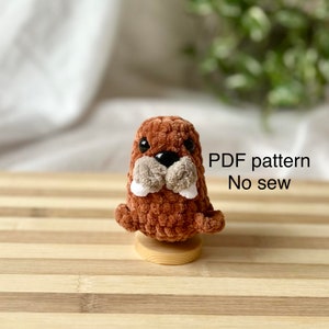Puede incluir: Un juguete de amigurumi de morsa de crochet marrón con colmillos blancos y ojos negros. El juguete está sentado sobre una base de madera. Patrón PDF Sin coser.