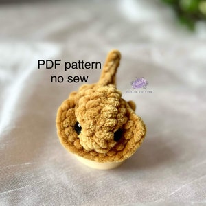 Puede incluir: Un juguete de amigurumi de raya de color marrón tejido a crochet con ojos negros. El juguete está hecho con una técnica sin costura y está disponible como patrón PDF. El texto "PDF pattern no sew" está escrito en blanco sobre fondo negro. El texto "Doux Coton" está escrito en morado sobre fondo blanco.