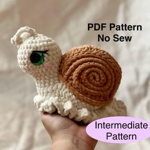 May include: A crocheted snail toy in cream and brown. The snail has large green eyes, pink cheeks, and a spiral shell. The text "PDF Pattern No Sew" and "Intermediate Pattern" are visible. The snail is held in a hand.