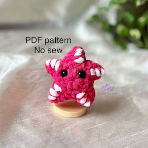 Puede incluir: Una estrella de mar de crochet rosa y blanca con ojos negros. El texto "PDF pattern No sew" está por encima de la estrella de mar.