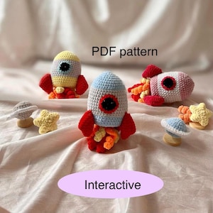 Puede incluir: Un conjunto de tres juguetes cohete de crochet con elementos interactivos, incluyendo un planeta y una estrella. Los cohetes son en tonos de azul, amarillo y rosa, con llamas rojas. El texto "PDF pattern" e "Interactive" es visible en la imagen.