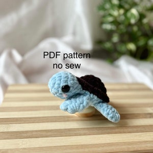 Może przedstawiać: Pluszowa zabawka amigurumi w kształcie żółwia z niebieskim ciałem, czarnym pancerzem i czarnymi oczami. Zabawka siedzi na drewnianej powierzchni. Wzór PDF bez szycia.