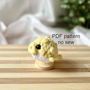 Peut inclure: Un jouet amigurumi en forme de poisson-globe crocheté en jaune et blanc avec des yeux noirs. Le jouet est posé sur une surface en bois. Patron PDF sans couture.
