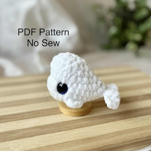 Può includere: Una balena bianca all'uncinetto con un occhio blu scuro, appoggiata su un piccolo supporto di legno. La balena ha un aspetto strutturato e soffice. Il testo "PDF Pattern No Sew" è visualizzato sopra la balena.