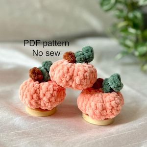 May include: Three crocheted pumpkins with green and brown stems. The pumpkins are a light peach color and are sitting on a white surface. The text "PDF pattern No sew" is visible in the image.