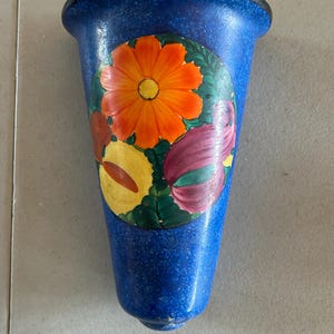 May include: A blue ceramic wall pocket vase with a speckled finish. The vase features a hand-painted floral design with orange, yellow, and purple flowers. The rim is trimmed with a black border.