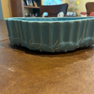 May include: A light blue ceramic bowl with a glossy finish. The exterior features a vertical ribbed pattern along the upper section and a decorative band of sculpted leaves encircling the base. The bowl has an oval shape with a wide opening.