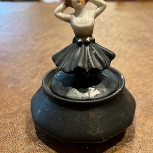 May include: A vintage black ceramic trinket box with a lid, topped with a small porcelain figurine of a woman with her hands behind her head. The figurine is wearing a black skirt and a white top.