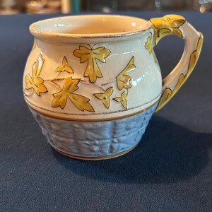 May include: A ceramic mug with a light blue, textured base and a cream-colored upper section decorated with yellow leaf designs. The handle is yellow with a decorative design. The mug is likely for beverages.