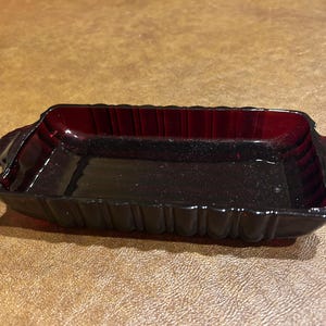 May include: A rectangular, deep red glass dish featuring a scalloped edge design and small handles on each side. The glass has a translucent quality with vertical ribbed detailing along the exterior walls and a smooth, flat interior base.