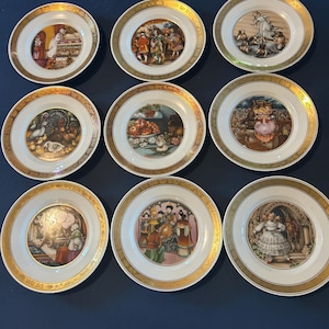 May include: Nine decorative porcelain plates with gold rims. Each plate features a different colorful illustration, depicting various scenes. The plates are arranged in three rows of three on a dark blue surface.