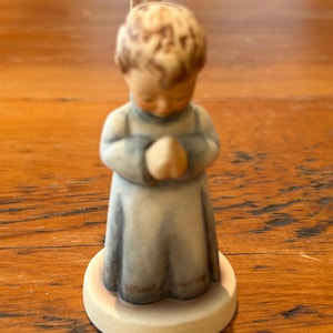 May include: A ceramic figurine of a child in a light blue robe with hands clasped in prayer. The figurine has a tan base and a detailed face with brown hair. The figurine is a religious item.