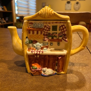 May include: A yellow ceramic teapot shaped like a kitchen scene. The front features a detailed relief of a counter, cabinets, a window with checkered curtains, a small white cat on the floor, and various kitchen items in brown, white, and blue.