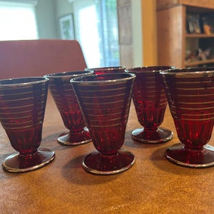 May include: A set of five ruby red glass footed tumblers, each featuring horizontal silver bands around the exterior. The glasses have a flared rim, a tapered body, and a circular base, all accented with silver trim along the edges and rims.