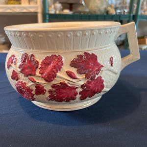 May include: Cream-colored ceramic chamber pot with a handle and embossed floral design. The pot features a band of small, raised dots around the rim and is decorated with red and maroon flowers.