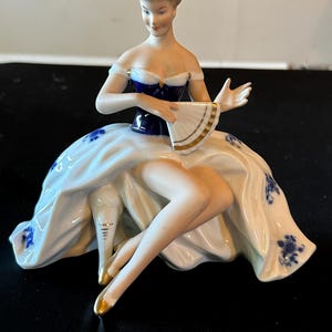 May include: A porcelain figurine of a seated figure wearing a dark blue bodice and a white skirt with blue floral patterns. The figure holds a white fan with gold trim and wears gold-tipped shoes, with hair styled in an updo and off-shoulder sleeves.