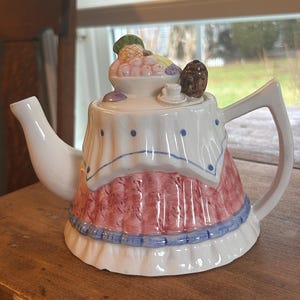 May include: A decorative ceramic teapot with a white base, a red and white patterned skirt, and a lid adorned with fruit and a small brown figurine. The teapot has a white spout and handle, and is sitting on a wooden surface.