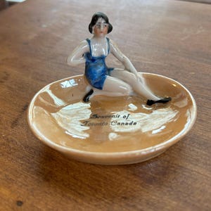 May include: A vintage ceramic dish with a seated figurine in a blue swimsuit. The dish is peach-colored with the text "Souvenir of Toronto Canada". The figurine has black hair and shoes.